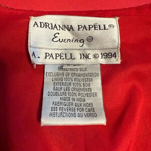 May include: A close-up of a red garment with a white tag. The tag reads "ADRIANNA PAPELL Evening" and "A. PAPELL INC &copy; 1994". The tag also lists the shell and lining materials.