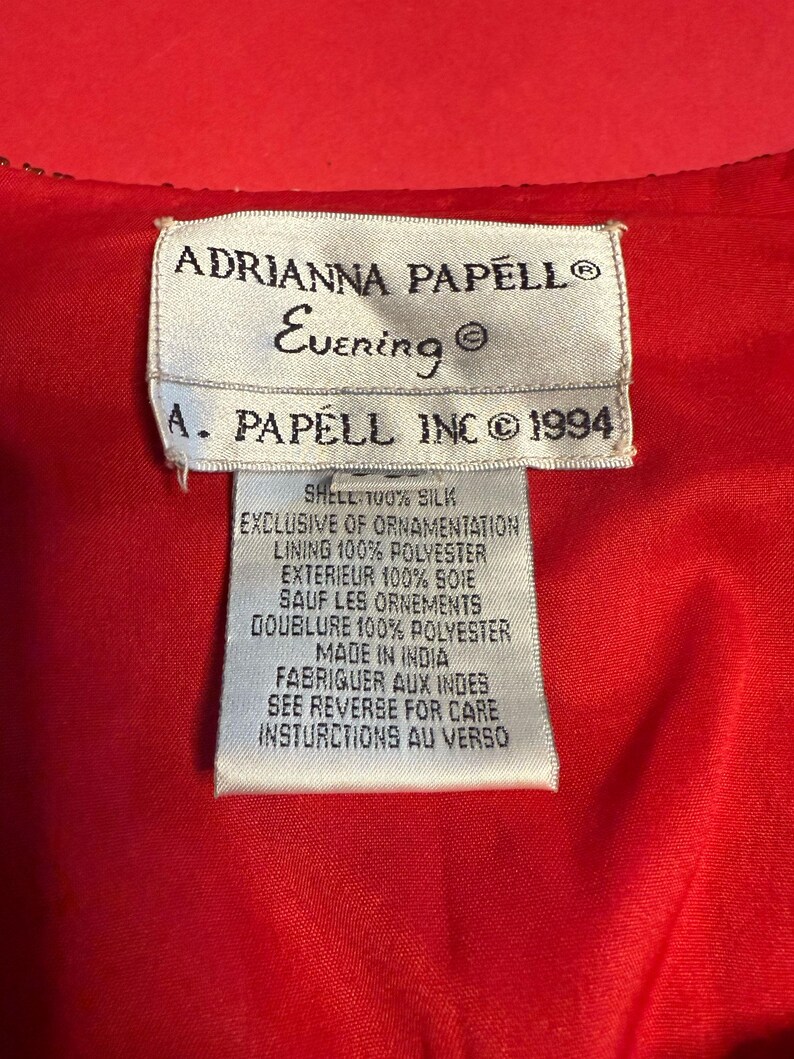 May include: A close-up of a red garment with a white tag. The tag reads "ADRIANNA PAPELL Evening" and "A. PAPELL INC &copy; 1994". The tag also lists the shell and lining materials.