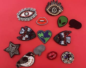 May include: Assortment of colorful embroidered iron-on patches featuring various designs, including eyes, lips, a heart, a star, a bird, a fish, an alien, a speech bubble, and a circle with a design inside.