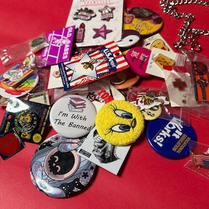 May include: A collection of colorful pins and badges on a red surface. The pins feature various designs, including text, cartoon characters, and patriotic themes. Some pins are packaged, and a silver chain is visible.
