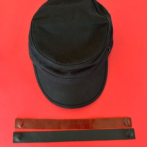 May include: A black canvas cap with a brown leather strap and a black leather strap with metal snaps.