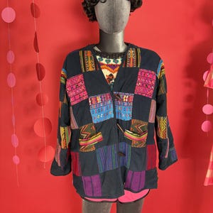 May include: A dark blue patchwork jacket with colorful embroidered squares. The jacket has a V-neck, two pockets, and wooden buttons. The mannequin is wearing a patterned sweater underneath and pink shorts.
