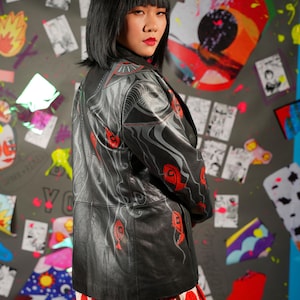 May include: A black leather jacket with a distinctive design in grey and red abstract patterns. The jacket is worn over a red and white polka dot skirt. The background includes colourful artwork and a person with black hair.
