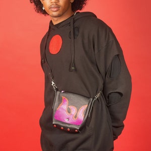 May include: A black hoodie with a red circle and holes in the sleeves. The person is wearing a red bucket hat with smiley faces and a black and purple crossbody bag with a flame design.