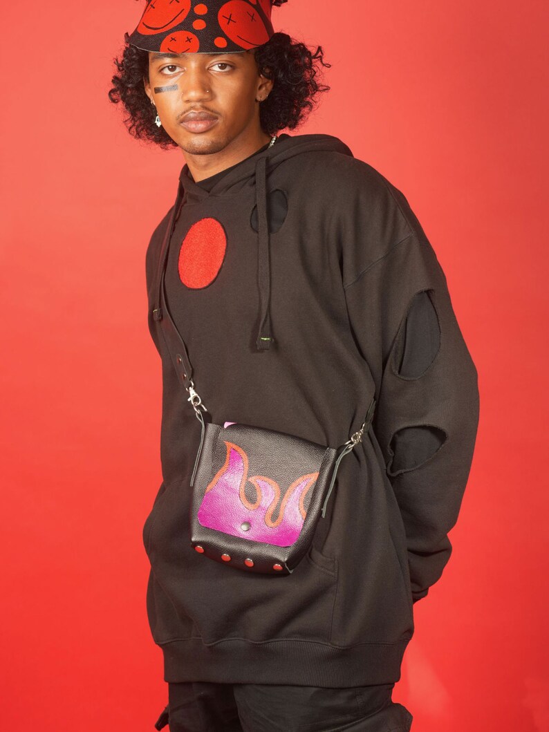 May include: A black hoodie with a red circle and holes in the sleeves. The person is wearing a red bucket hat with smiley faces and a black and purple crossbody bag with a flame design.