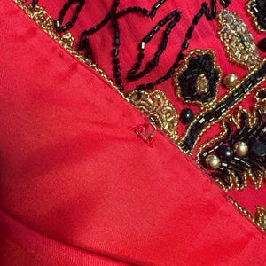 May include: Close-up of a red garment with intricate beadwork. The design features black and gold beads arranged in floral and abstract patterns. The fabric appears to be a satin or silk material.