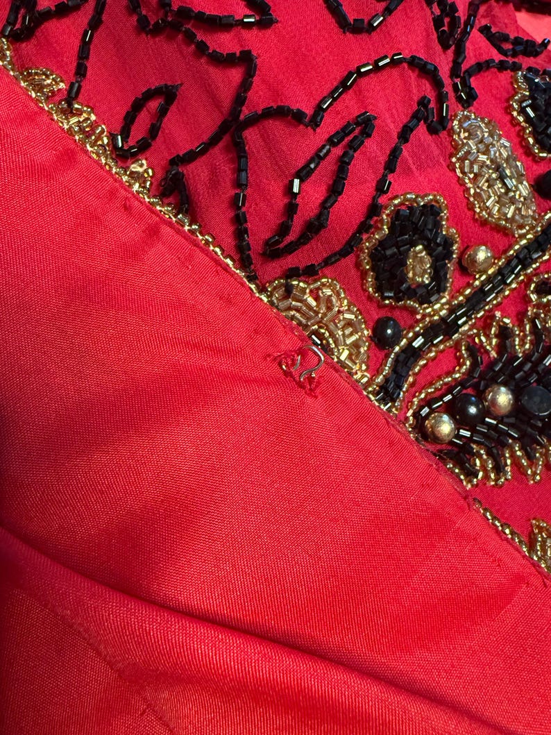May include: Close-up of a red garment with intricate beadwork. The design features black and gold beads arranged in floral and abstract patterns. The fabric appears to be a satin or silk material.