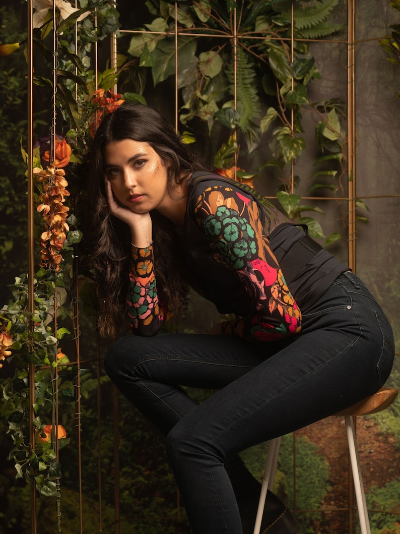 May include: A woman wearing a black top with a colorful floral print and dark blue jeans sits on a stool in front of a green leafy backdrop.