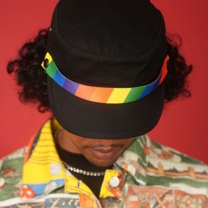 May include: A black military style cap with a rainbow colored band around the crown. The cap is worn by a person with curly hair and a colorful patterned shirt.