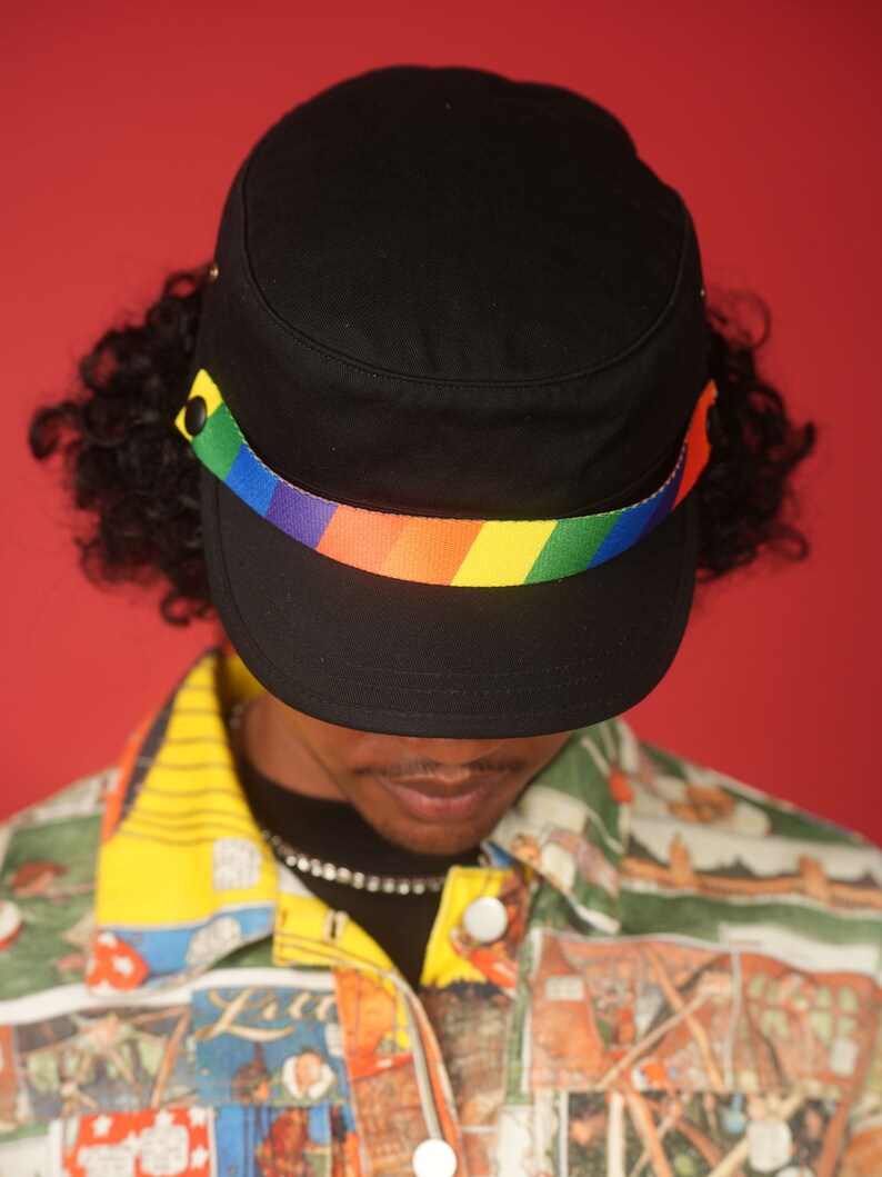 May include: A black military style cap with a rainbow colored band around the crown. The cap is worn by a person with curly hair and a colorful patterned shirt.