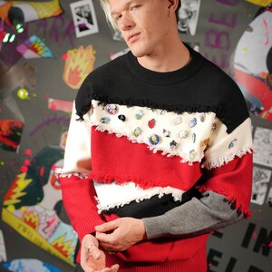 May include: A multi-coloured sweater with a black base and a red, white, and grey patchwork design. The sweater has a frayed edge and is decorated with various pins.