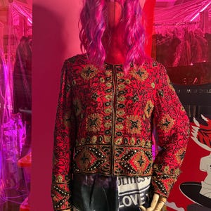 May include: A red jacket with intricate gold and black embroidery, displayed on a mannequin with a pink wig. The jacket features a floral pattern and beaded accents. The bottom of the mannequin is wearing a skirt with the word "LOVE" printed on it.