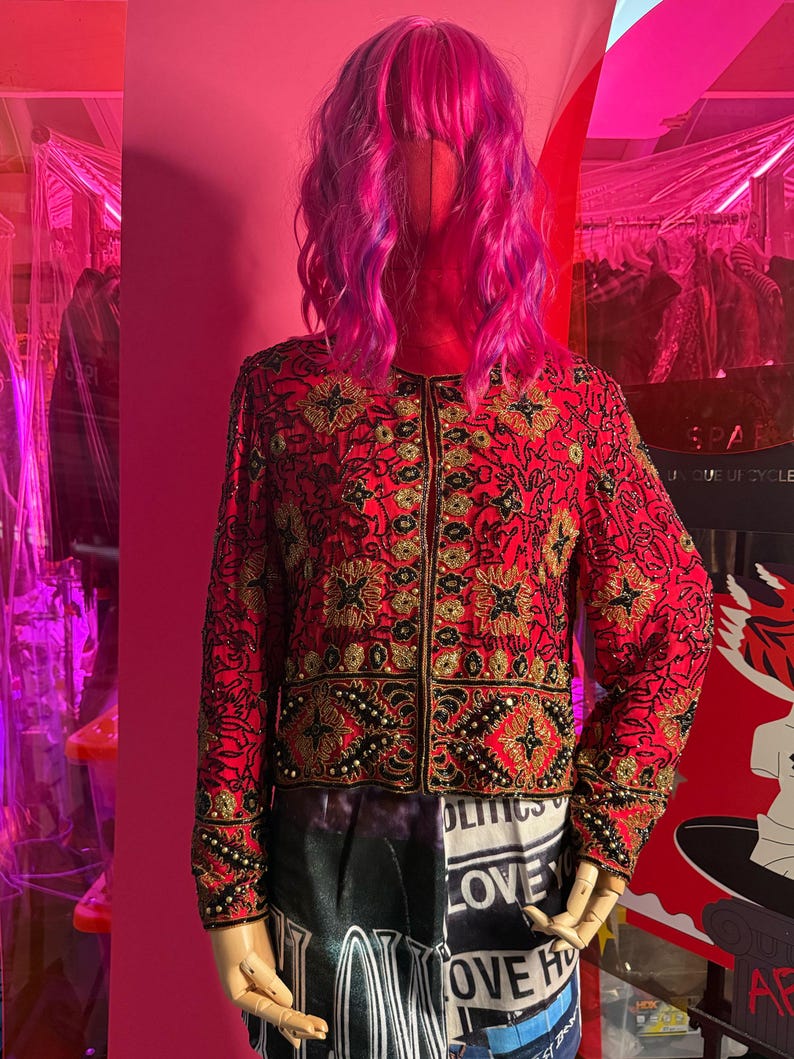May include: A red jacket with intricate gold and black embroidery, displayed on a mannequin with a pink wig. The jacket features a floral pattern and beaded accents. The bottom of the mannequin is wearing a skirt with the word "LOVE" printed on it.