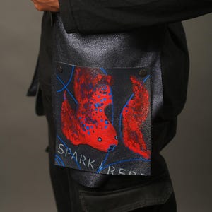 Puede incluir: Una bolsa de cuero negra con un diseño de pez rojo y azul y el texto "SPARK FIRE" impreso en la parte delantera.