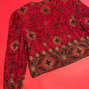 May include: A red jacket with intricate black and gold beadwork. The jacket features a floral pattern and a decorative border along the hem and cuffs. The sleeves are long, and the jacket is fully embellished.