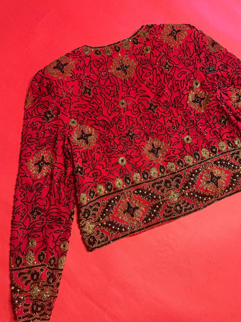 May include: A red jacket with intricate black and gold beadwork. The jacket features a floral pattern and a decorative border along the hem and cuffs. The sleeves are long, and the jacket is fully embellished.