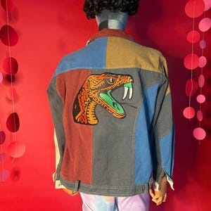 May include: A patchwork denim jacket with a large snake patch on the back. The jacket features panels of blue, brown, red, and grey denim. The snake patch is orange, green, and black.