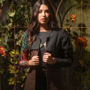 May include: A woman wearing a brown and black long-sleeved jacket with a floral print on the sleeves. The jacket has a zipper closure and a collar. The woman is standing in front of a green leafy background.