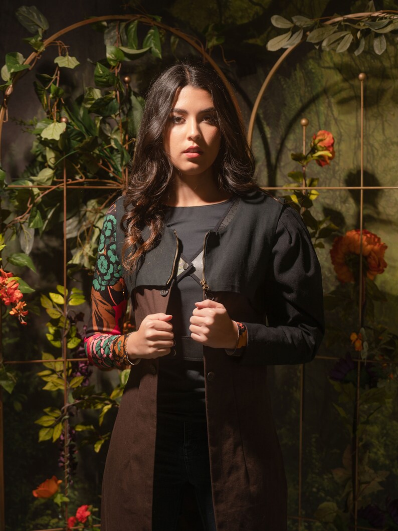 May include: A woman wearing a brown and black long-sleeved jacket with a floral print on the sleeves. The jacket has a zipper closure and a collar. The woman is standing in front of a green leafy background.