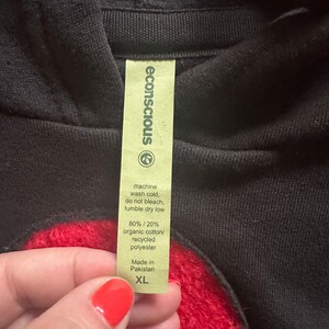 May include: Black hoodie with a red heart design and a green tag that says "econscious" and includes care instructions and fabric content. The tag also says "Made in Pakistan" and "XL".