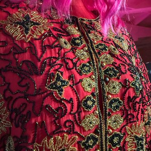 May include: A red jacket with intricate gold and black beaded floral embroidery. The jacket features a high collar and a central zipper. The design includes gold beads and black accents, creating a detailed and ornate pattern.