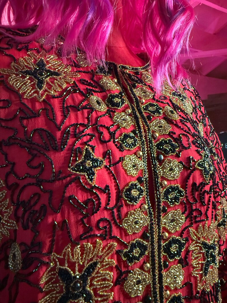 May include: A red jacket with intricate gold and black beaded floral embroidery. The jacket features a high collar and a central zipper. The design includes gold beads and black accents, creating a detailed and ornate pattern.