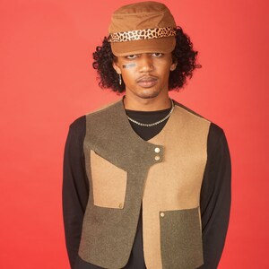 May include: A person wearing a brown and black patterned cap, a black long-sleeved shirt, and a brown and green vest with multiple pockets and gold buttons.