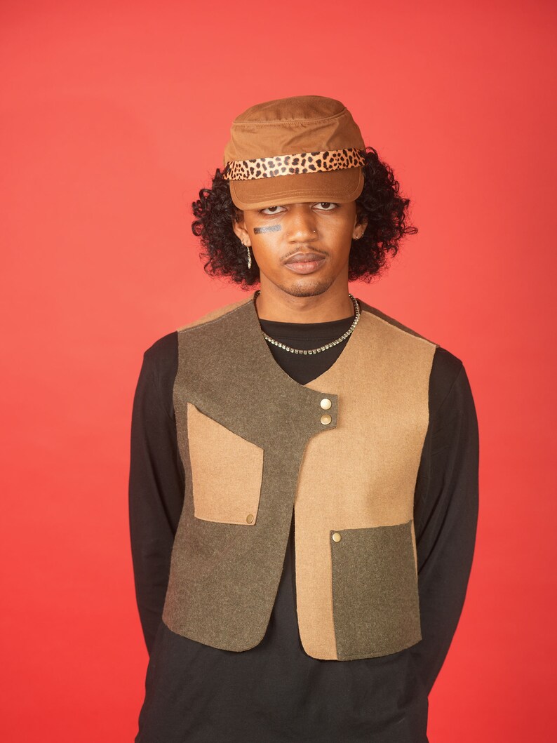 May include: A person wearing a brown and black patterned cap, a black long-sleeved shirt, and a brown and green vest with multiple pockets and gold buttons.