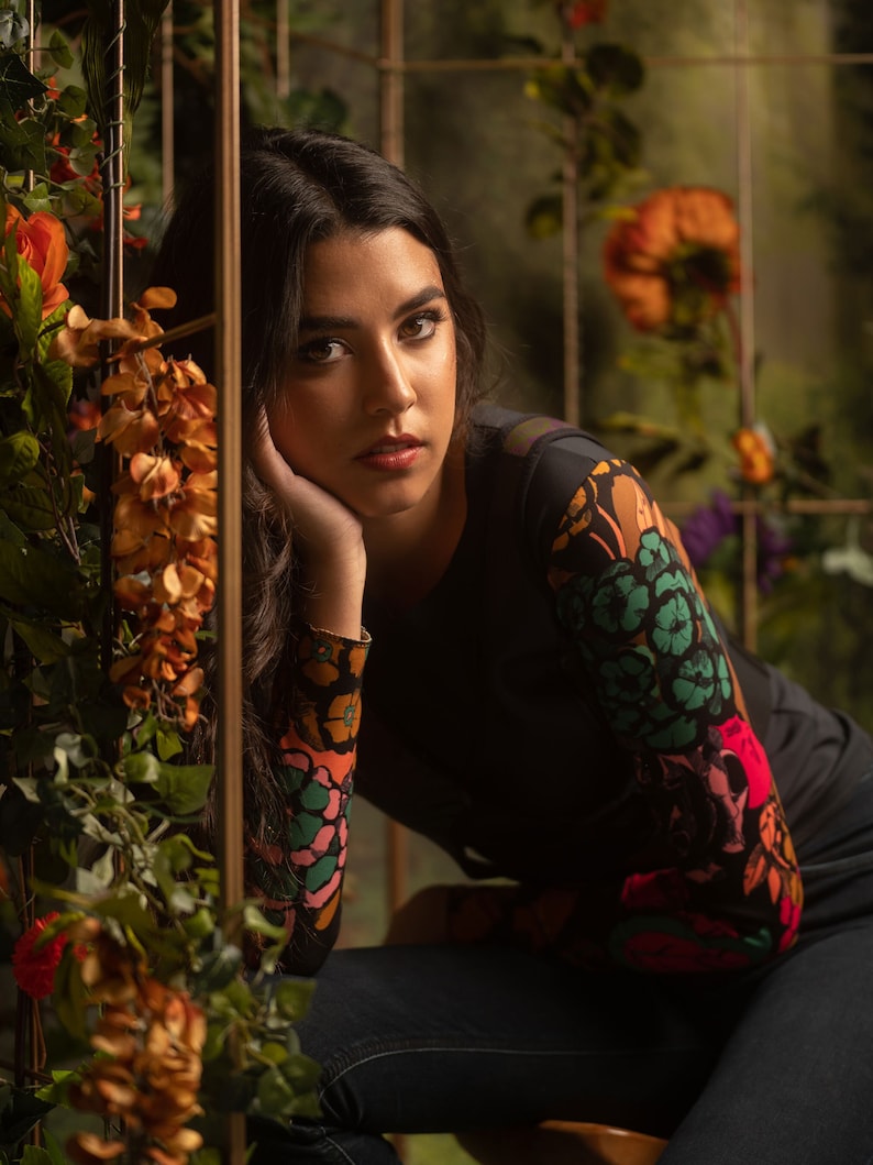 May include: A woman with long dark hair is wearing a black long-sleeved shirt with a colorful floral print. She is sitting with her chin resting on her hand and looking at the camera.