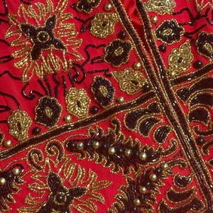 May include: Close-up of a red garment with intricate gold and black beadwork. The design features floral and geometric patterns, with a central vertical seam. The beads are arranged in detailed floral and leaf motifs.