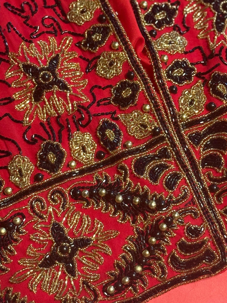 May include: Close-up of a red garment with intricate gold and black beadwork. The design features floral and geometric patterns, with a central vertical seam. The beads are arranged in detailed floral and leaf motifs.