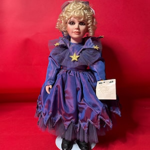 Könnte beinhalten: Porzellanpuppe mit blonden Locken, trägt ein blau-violettes Kleid mit Sternen. Die Puppe hat ein detailliertes Gesicht und hält ein Etikett mit der Aufschrift "Designer Guild". Die Puppe steht auf einem weißen Sockel.
