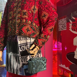 May include: A red jacket with gold and black embroidery, paired with a black and white printed skirt and red pants. A small, teal beaded purse hangs from a chain. The outfit is displayed on a mannequin.