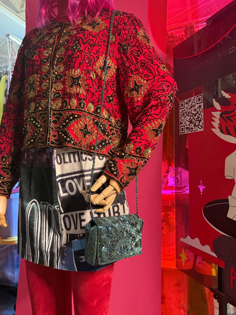May include: A red jacket with gold and black embroidery, paired with a black and white printed skirt and red pants. A small, teal beaded purse hangs from a chain. The outfit is displayed on a mannequin.