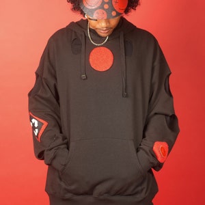 May include: A black hoodie with red patches and cutouts. The hoodie has a large red circle on the front and a smaller red patch on the left sleeve. The person is wearing a black visor with red smiley faces and X's.