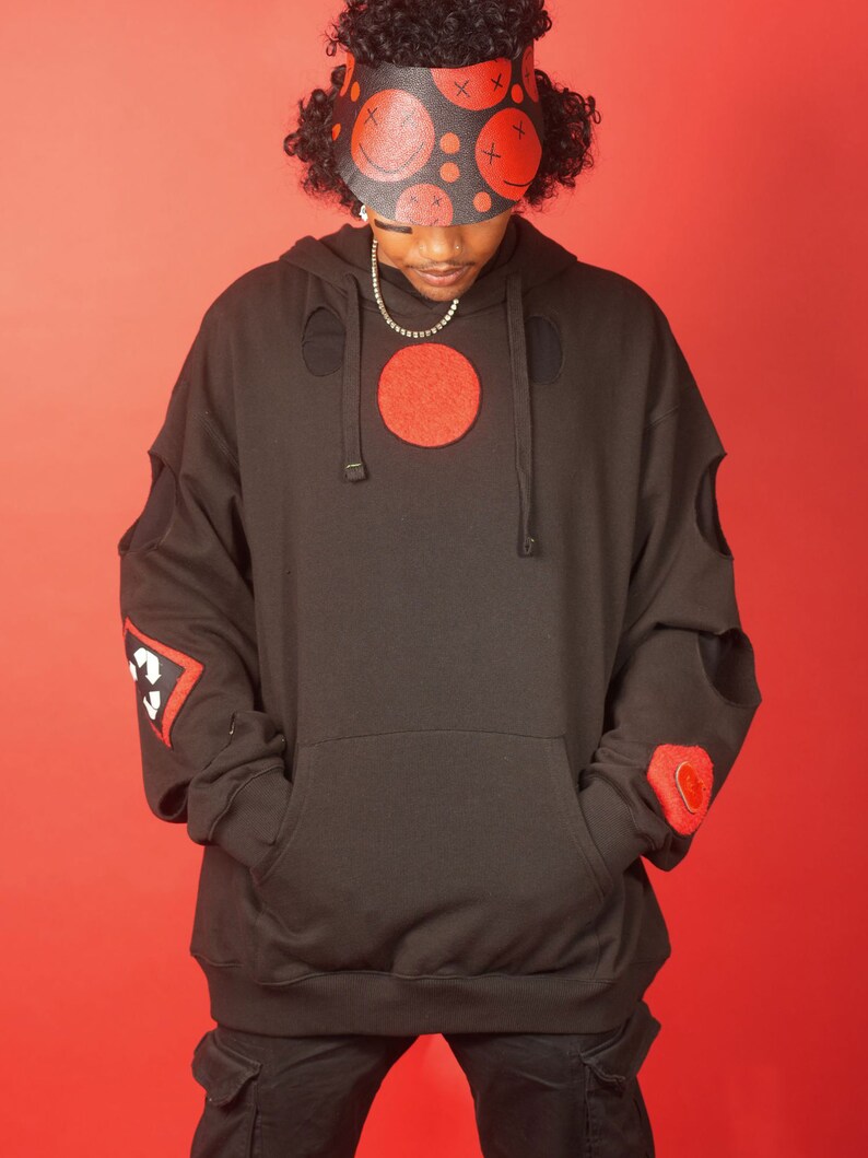 May include: A black hoodie with red patches and cutouts. The hoodie has a large red circle on the front and a smaller red patch on the left sleeve. The person is wearing a black visor with red smiley faces and X's.