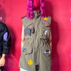 May include: An olive green vest with multiple patches, including a skull and crossbones, a smiley face, and a lightning bolt. The vest is worn over a light green skirt. A mannequin head is covered in pink hair.