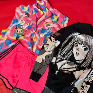 Op de afbeelding: Een verzameling kleurrijke kledingstukken op een rode achtergrond. Bestaat uit een roze en blauw vest met patronen en spelden, een felroze sportbroek met zwarte strepen en een zwarte trui met een manga-achtige afbeelding.