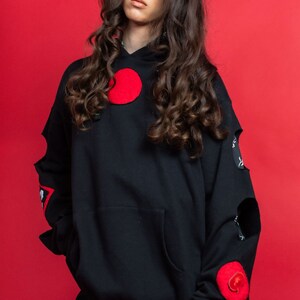 May include: A black hoodie with red circular patches and distressed detailing. The hoodie has a large pocket on the front and a drawstring hood.