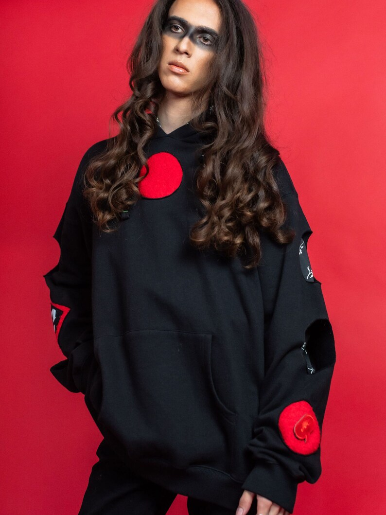 May include: A black hoodie with red circular patches and distressed detailing. The hoodie has a large pocket on the front and a drawstring hood.