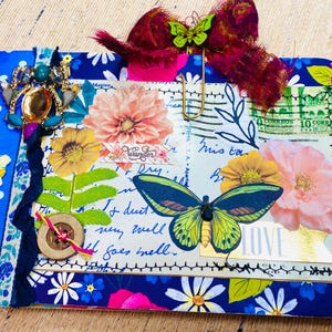 May include: A blue and white fabric covered journal with a variety of embellishments including a butterfly, flowers, and a jeweled spider. The journal has a handwritten message that reads "It doesn't run well, but it goes well".