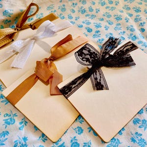 May include: Three blank cards with different colored ribbons tied around them. The ribbons are white, brown, and black lace. The cards are on a blue and white floral fabric.