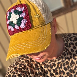 May include: A mustard yellow baseball cap featuring a square crochet patch on the front panel. The patch displays a floral pattern in white, dark green, and light purple. The cap has a beige mesh back and a distressed brim with frayed edges.