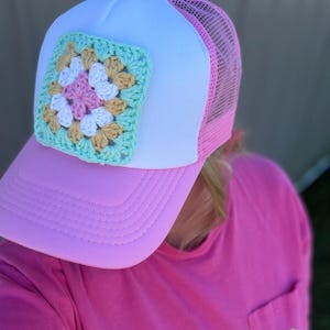 May include: A trucker hat featuring a white front panel, a pink mesh back, and a pink brim. A square crochet granny square patch in shades of mint green, white, tan, and light pink is centered on the front white panel of the hat.