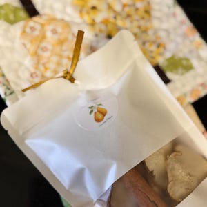 May include: A white semi-transparent pouch tied with a gold ribbon, featuring a circular sticker with a pear illustration and the text "Simmering Pears". Inside the pouch are visible dried pear slices and a brown cinnamon stick, resting on a quilted fabric.