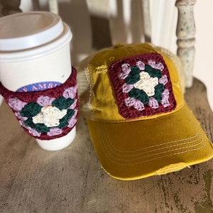 May include: A mustard yellow baseball cap with a distressed brim and a mesh back, featuring a square crochet granny square patch in maroon, dark green, and cream. Beside it is a white coffee cup with a matching crochet sleeve.