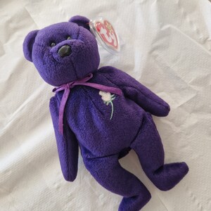 Puede incluir: Un oso de peluche morado con una nariz negra y una rosa blanca bordada en el pecho. El oso tiene una cinta morada alrededor del cuello y una etiqueta roja y blanca en la oreja.