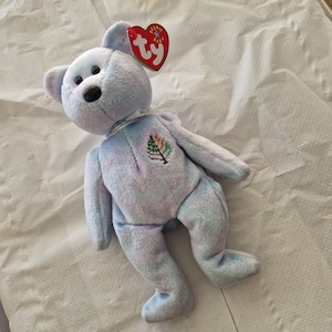 以下が含まれることがあります： 胸に小さな刺繍の木の模様が入った、水色のぬいぐるみ。クマの耳には赤いハート型のタグが付いています。クマは白い面に横たわっています。
