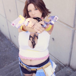 Pode incluir: Uma mulher com um traje branco e amarelo segura duas pistolas de brinquedo roxas e prateadas. Ela está usando uma saia azul e branca e um cinto marrom com franjas brancas.