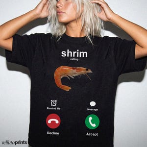 A woman wearing a black t-shirt with a graphic design depicting a shrimp and various icons representing different phone functions, such as a reminder, message, decline, and accept.